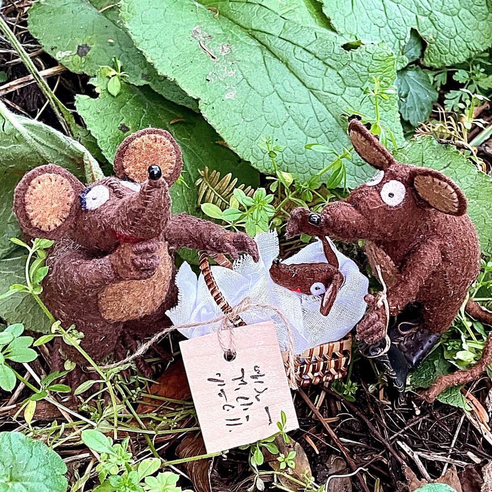 Photo of Minimus and Minima, the Latin mice, in a clump of grass and leaves. There's a wicker basket between them, with a label tied to it. In the basket is a smaller mouse, wrapped in a cloth