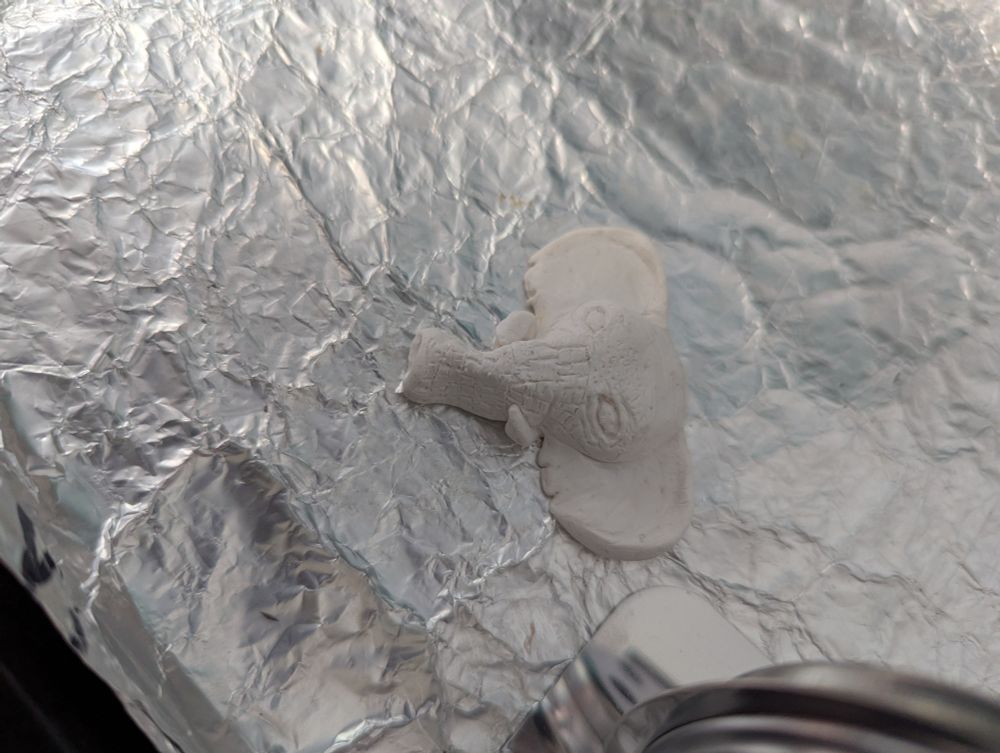 Side view of a white clay elephant sitting on some tinfoil next to an oven thermometer 