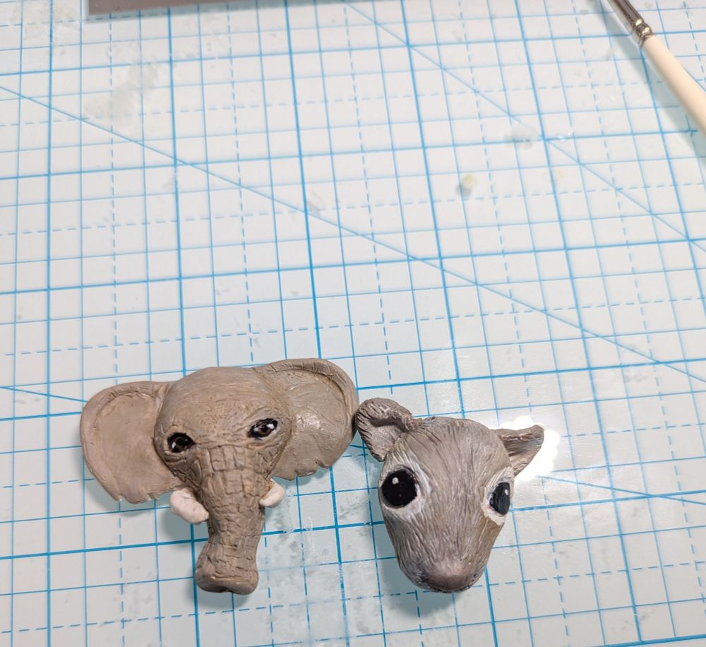 A little squirrel head and an elephant head made of polymer clay sit on a glass grid board. A paintbrush is up in the top right corner of the frame