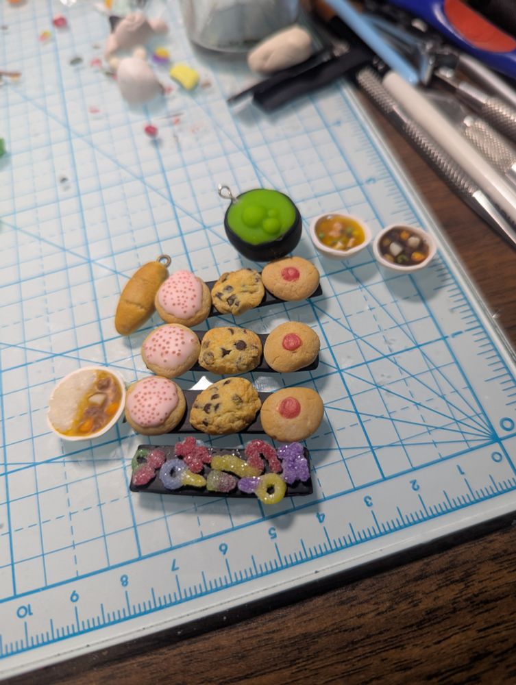 A bunch of unvarnished polymer clay miniatures, including a cauldron with green bubbly goo, two bowls of soup (one beef stew and one chicken vegetable!) Three hair pin bases that are black clay rectangles each with with three cookies on them (an iced pink sugar cookie with sprinkles, a chocolate chip cookie and a jam cookie), a hair clip base with tiny sour candies all over it, a very small loaf of bread and a little bowl of japanese curry 