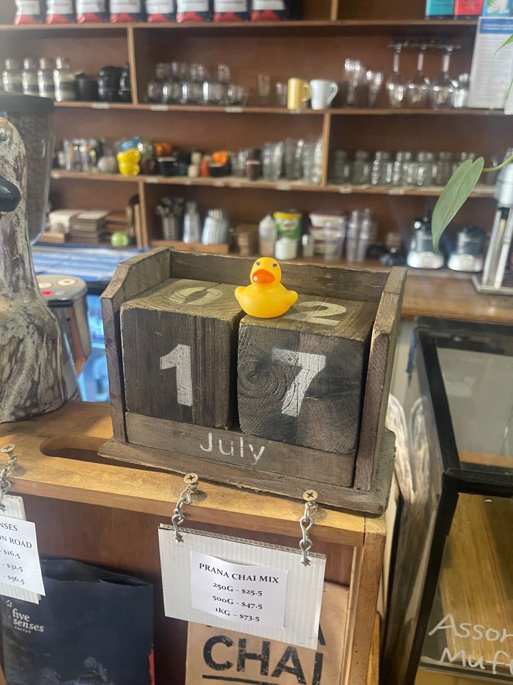 A small yellow rubber duck on a wooden calendar made of blocks. The calendar says “17 July”. The background of the photo is of a cafe.