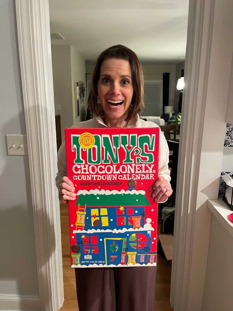 A middle aged woman with a brunette bob is standing in a doorway holding an absolutely gigantic Tony’s Chocolonely advent calendar and sporting an even more gigantic grin