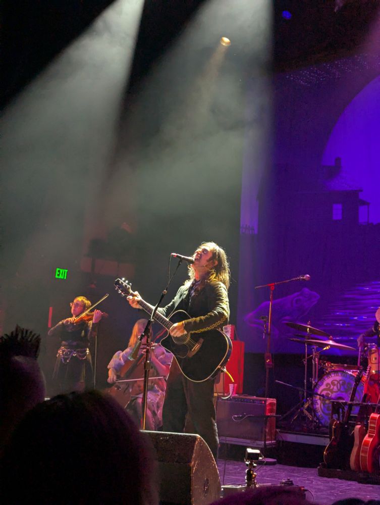 Pic from the show. A man with a guitar is singing in the spotlight, while a cellist and violinist play to the side behind him.