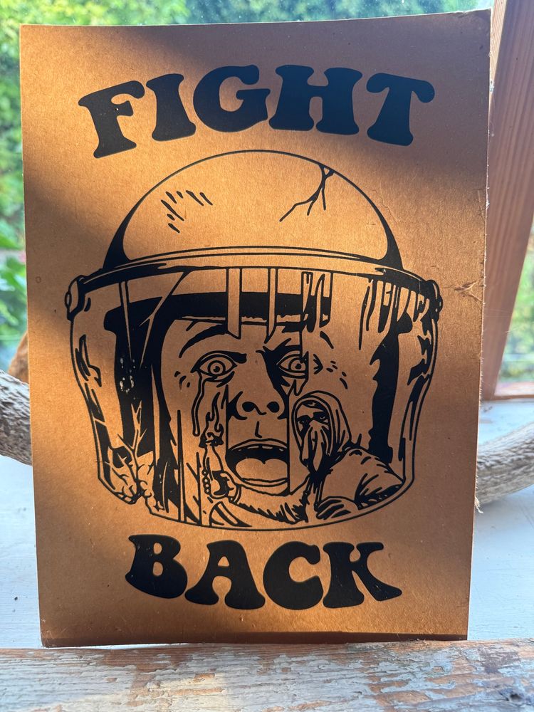 Ein Druck der einen behelmten Polizisten mit erschrecktem Gesichtsausdruck zeigt in dessen Spiegelung des Visiers eine vermummte Person mit einem brennenden Molotowcocktail zu sehen ist. Darüber und darunter die Worte "Fight" (oben) "back" (unten). Dies auf braunem dickem Papier.