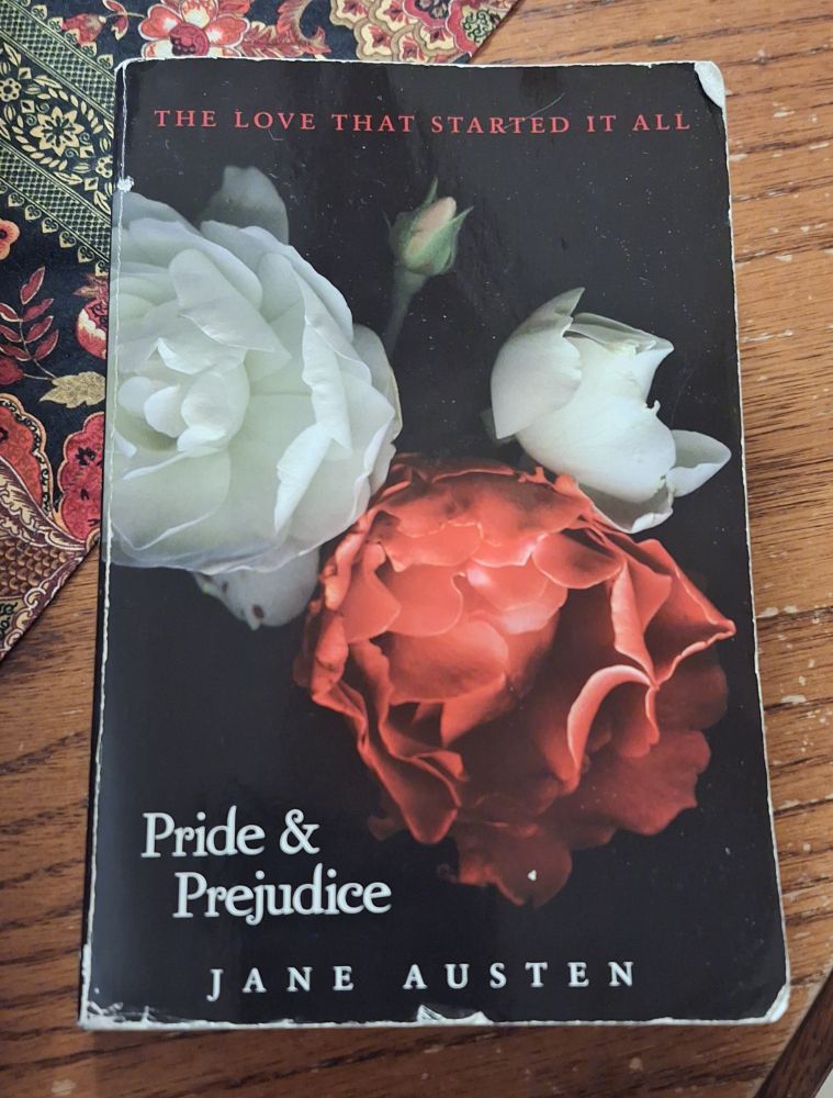 A copy of Jane Austen's novel "Pride and Prejudice" sitting on a wood table. Beneath the book is a dark colour patterned floral table runner. The cover is black in colour with red and white hydrangeas. The top of the cover has the line "the love that started it all" in red all caps lettering. "Pride & Prejudice" is written in white lettering, sitting to the bottom left of the flowers. "Jane Austen" is written in all caps white lettering. The cover is well loved.