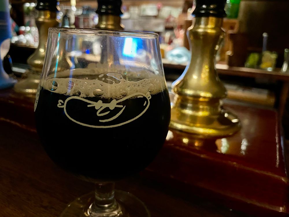 A goblet of dark beer with a light frothy head on a bar counter with handpumps.