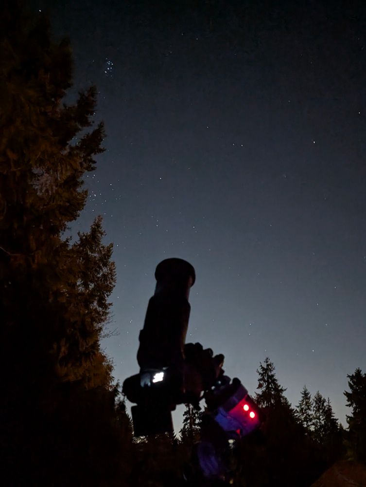 a mirrorless camera with a long lens mounted on a star tracker on a tripod, pointing at the sky. in the background are evergreens framing a night sky with a few stars visible; notably the Pleiades cluster is peeking out from the trees. 
