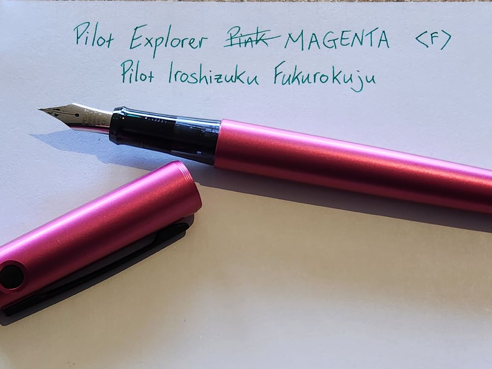 A pi- err magenta pilot explorer fountain pen, arranged on a sheet of paper identifying it as a pilot explorer fine nib, and the ink inside as Pilot Iroshizuku Fukurokuju green