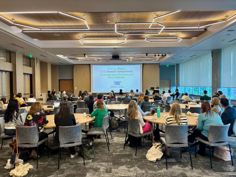 The Colorado Cancer Coalition Symposium being kicked off at the Elliman Conference Room at the Anschutz Health Sciences Building. There are many people sitting at round tables while watching the start of the presentation. 