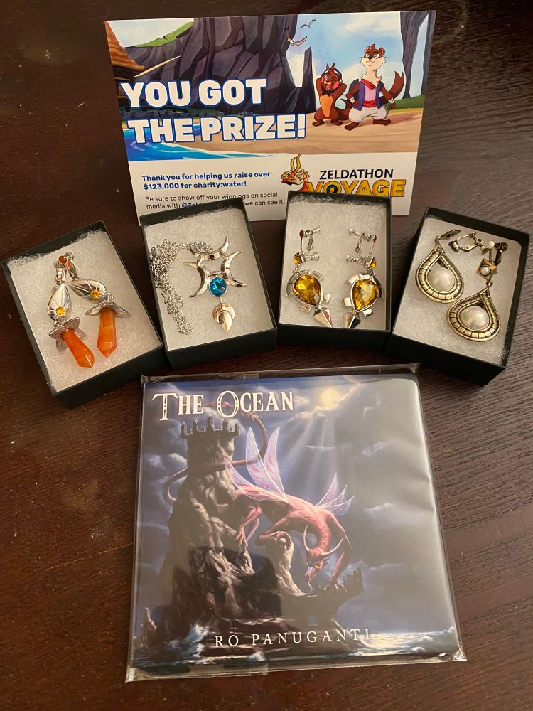 Photo showing three sets of earrings and a necklace based on Breath of thd Wild in front of a postcard saying “You got the prize!” From Zeldathon, and a CD called The Ocean by Ro Panuganti. 