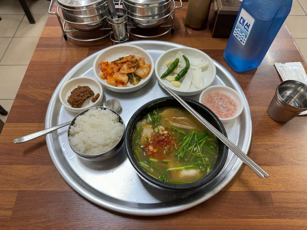 Eine Schüssel dwaejigukbap, eine Suppe mit Schweinefleisch und Reis. Daneben Schälchen mit natürlich Toppings und Kimchi