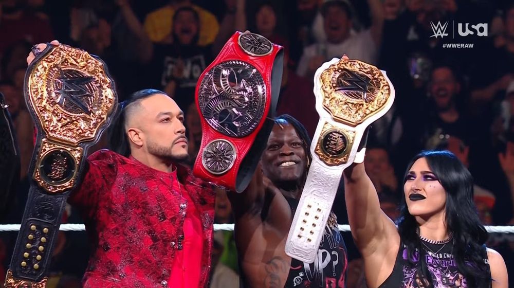 Damian Priest holding up the world heavyweight title, Rhea Ripley holding up the women’s world title with R-Truth in the middle of them holding up the RAW tag team championship. Damian and Rhea both looking at the tag team title in between in disbelief 