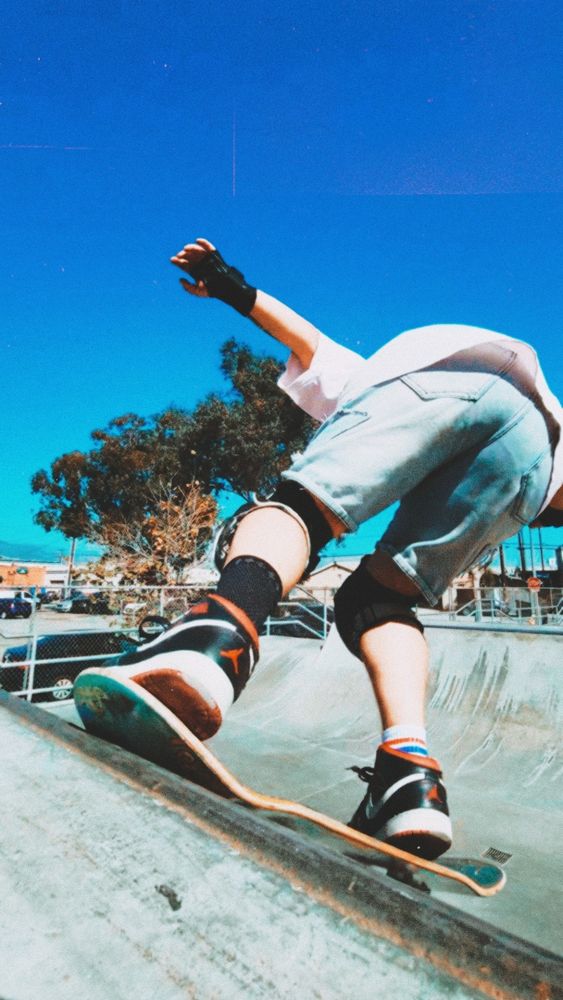 Ore is mid dropping in on a skateboard on a concrete halfpipe from a camera perspective of on the floor looking up. We only see the back of their jean shorts, knee pads, pink t-shirt and they have red and black sneakers on. The angle makes it so we cannot see their head.