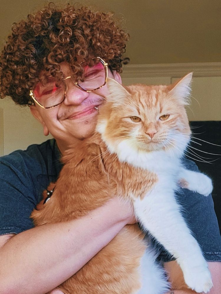  A selfie of Ore who has brown curly hair and brown chunky square glasses, holding a fluffy white and orange cat named Kickflip. She looks angry at the camera. 