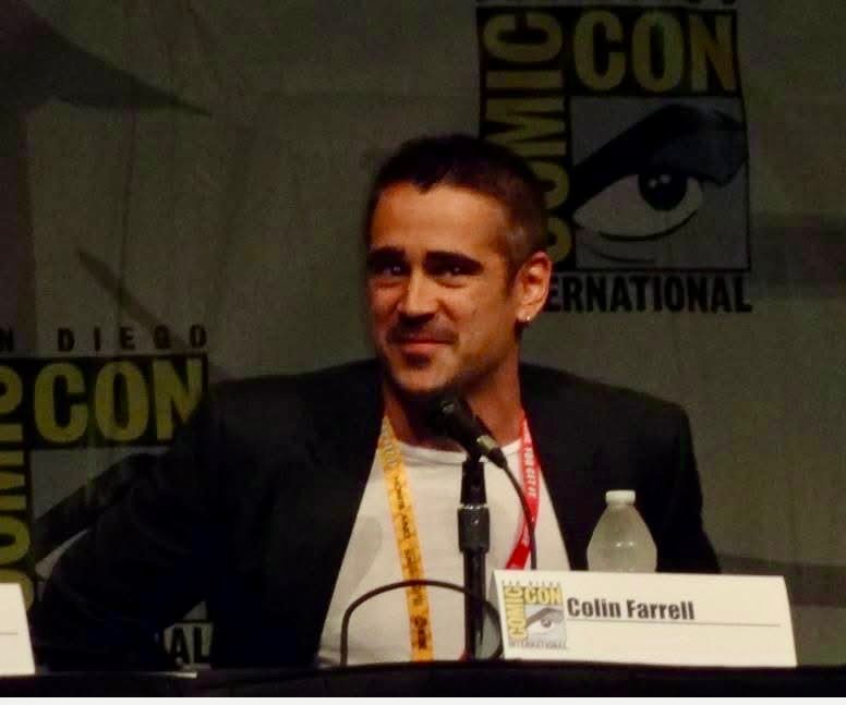 Actor Colin Farrell, a white dude with short dark hair and eyes, with bold eyebrows, wearing a white top and black blazer, on a San Diego Comic Con stage. He is smiling slightly. A card with his name and a bottle of water sit on the table in front of him. 