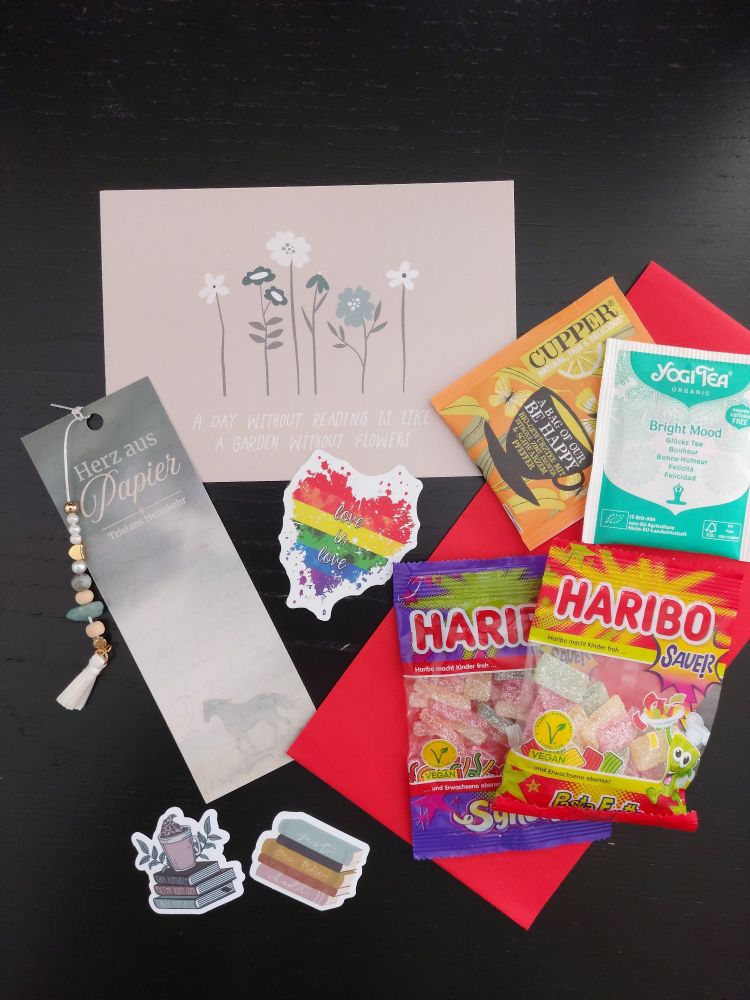 Goodies: Lesezeichen mit einer Schnur aus winzigen Steinen, Postkarte, Sticker, darunter ein Pride-Herz mit dem Text "Love is Love"  , Haribo und Tee.