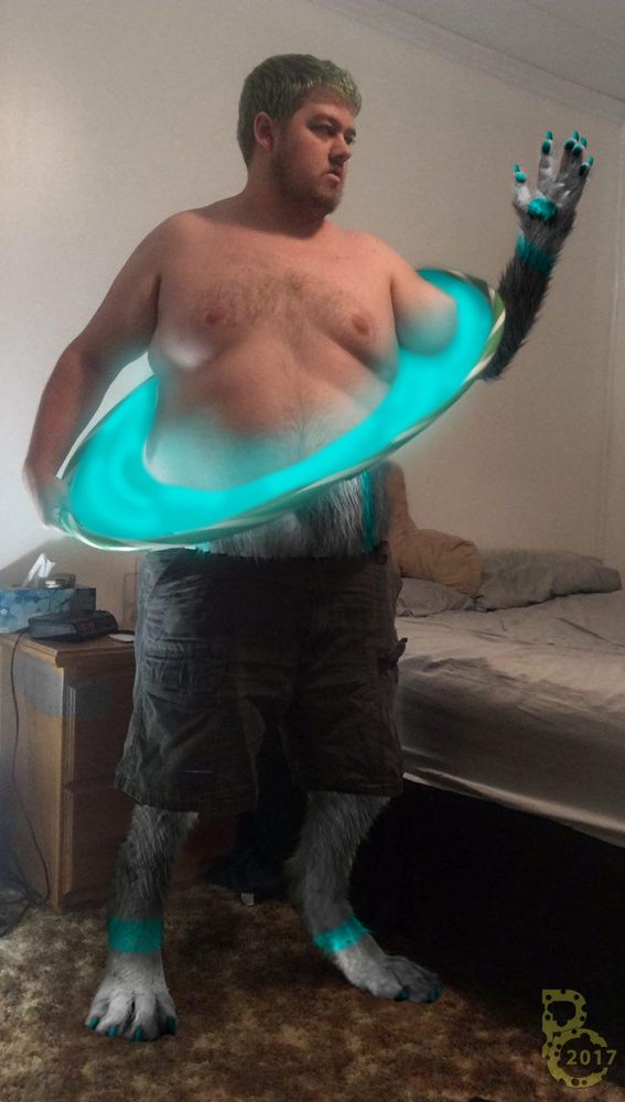 A photomanipulation of a large shirtless man in cargo shorts standing in a bedroom.  He is transforming into a canine furry with gray, white and teal fur from the feet up via a magic hoop, which is pulled up to his belly.  One forearm is looped through the hoop, and he is looking at the clawed, furred hand that is coming out of the other side.