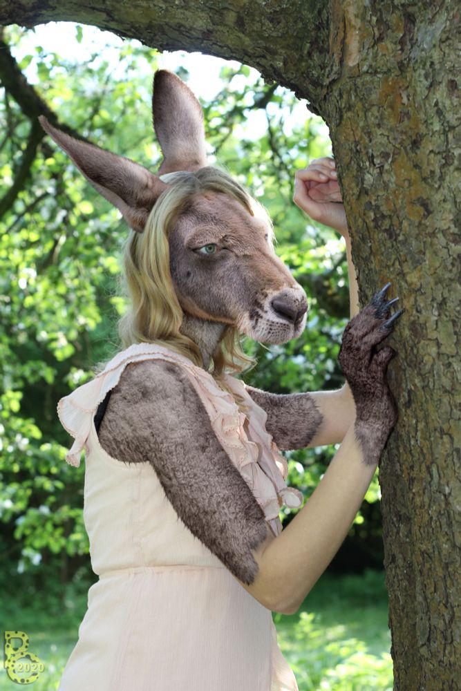 A photomanipulation of a medium shot of a blonde woman in a cream dress leaning against a tree as she transforms into an anthro kangaroo