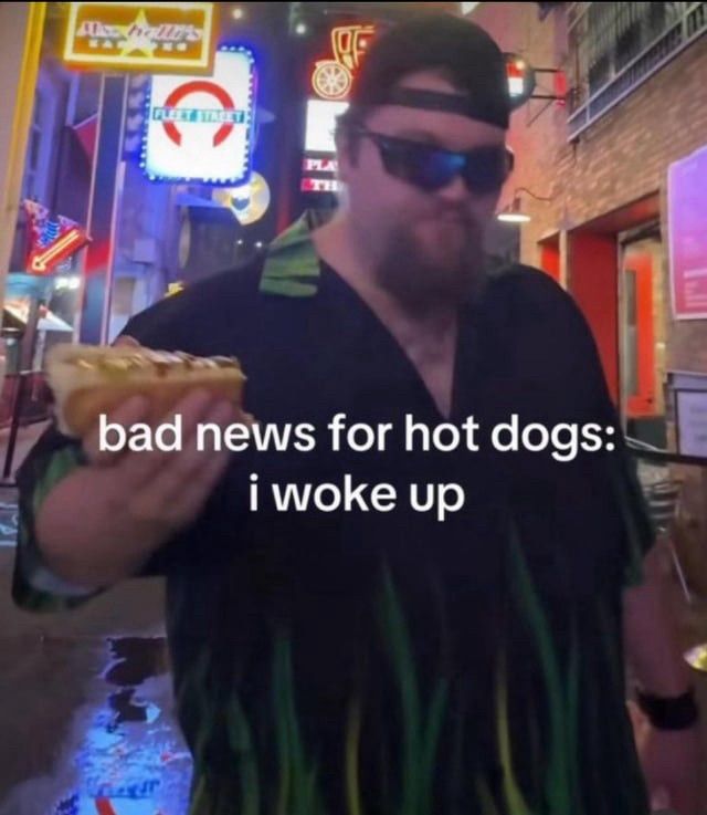 A big guy with a beard wearing shades, a backwards cap, and a black short sleeved button up shirt with what is maybe green flames or grass blades on it. He's standing on a sidewalk or in the street and holding a hotdog. He looks somewhat menacing. The caption reads: "Bad news for hot dogs: I woke up".