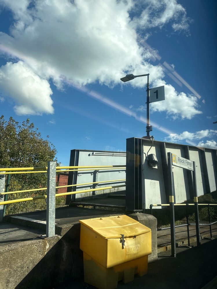 A picture showing the Rock Ferry platform sign in front of the rising walkway to cross platforms. A bright blue sky with a white fluffy cloud and on the platform there is a yellow salt bucket.