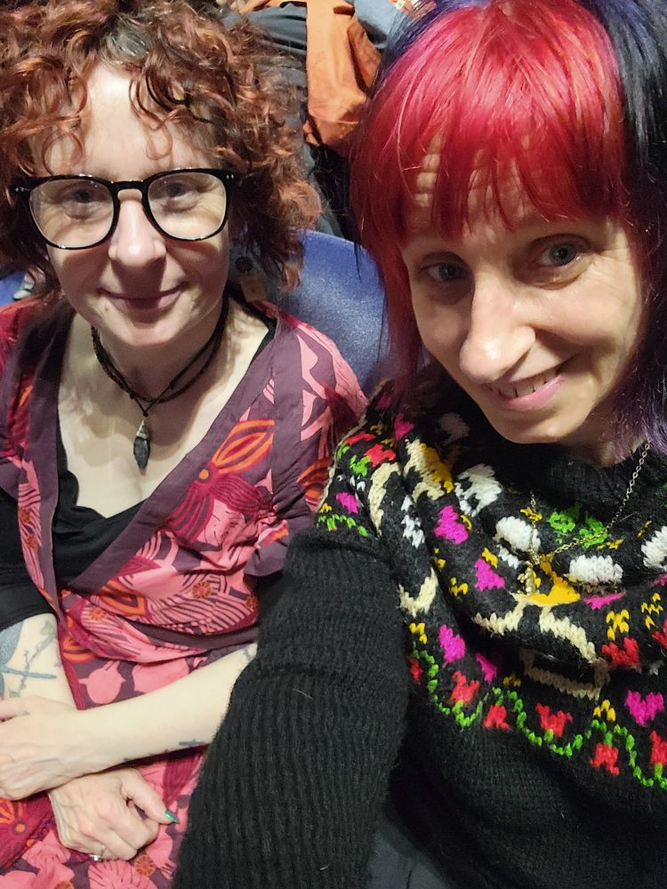 Two women seated in a theatre. Both are smiling and wearing colourful clothing.