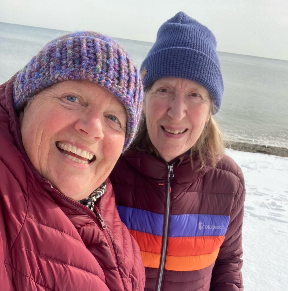 Selfie of two smiling white women, both are wearing maroon puffer jacket and purple hats. They are cousins. 