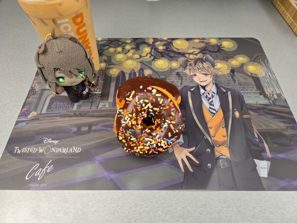 A photograph of two donuts, a cup of iced coffee, and a small plush of Leona Kingscholar from Twisted Wonderland. The items are arranged on a placemat from the Twisted Wonderland Café collaboration, featuring Ruggie Bucchi.