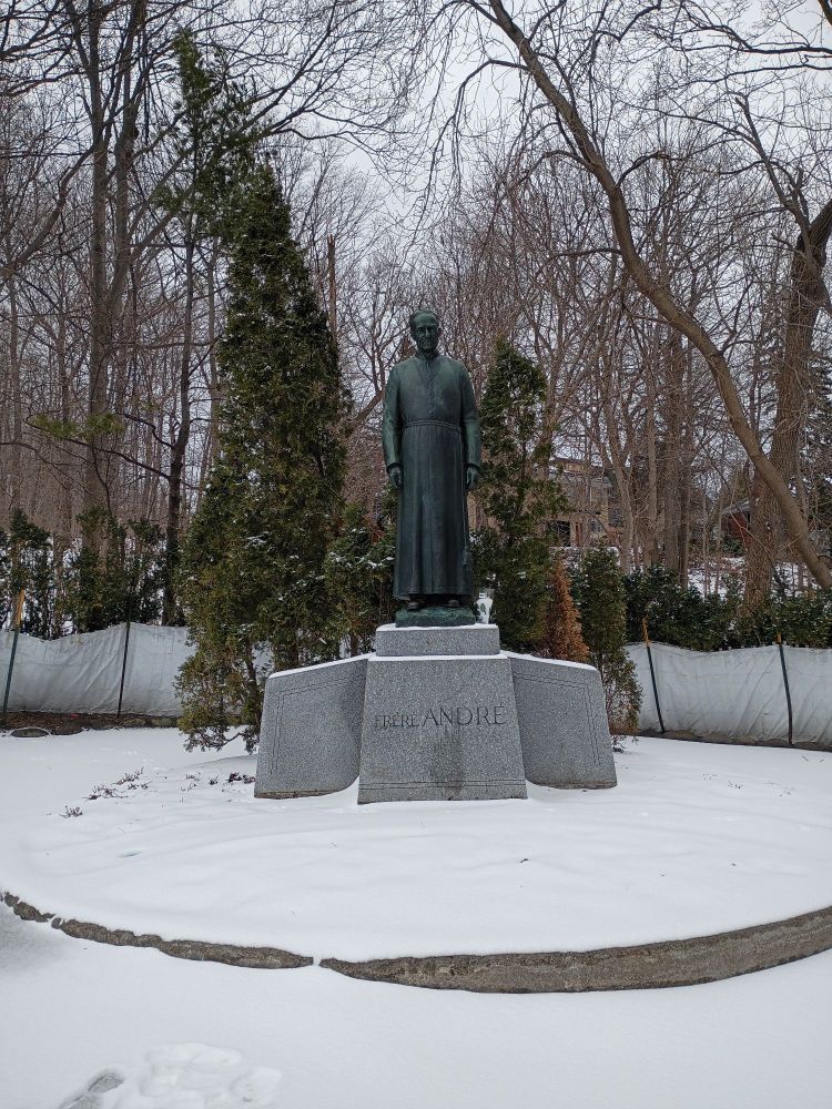 Statue of Saint Brother Andre Bessette set against a background of trees on a snowy day.