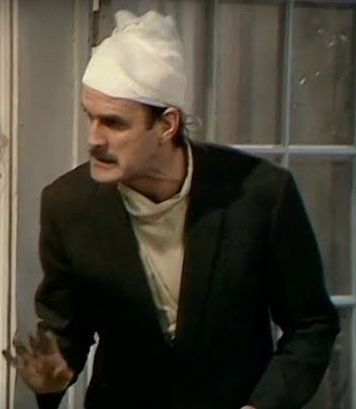 John Cleese as Basil Fawlty, head wrapped in a bandage, cautioning that German guests should be engaged with care 