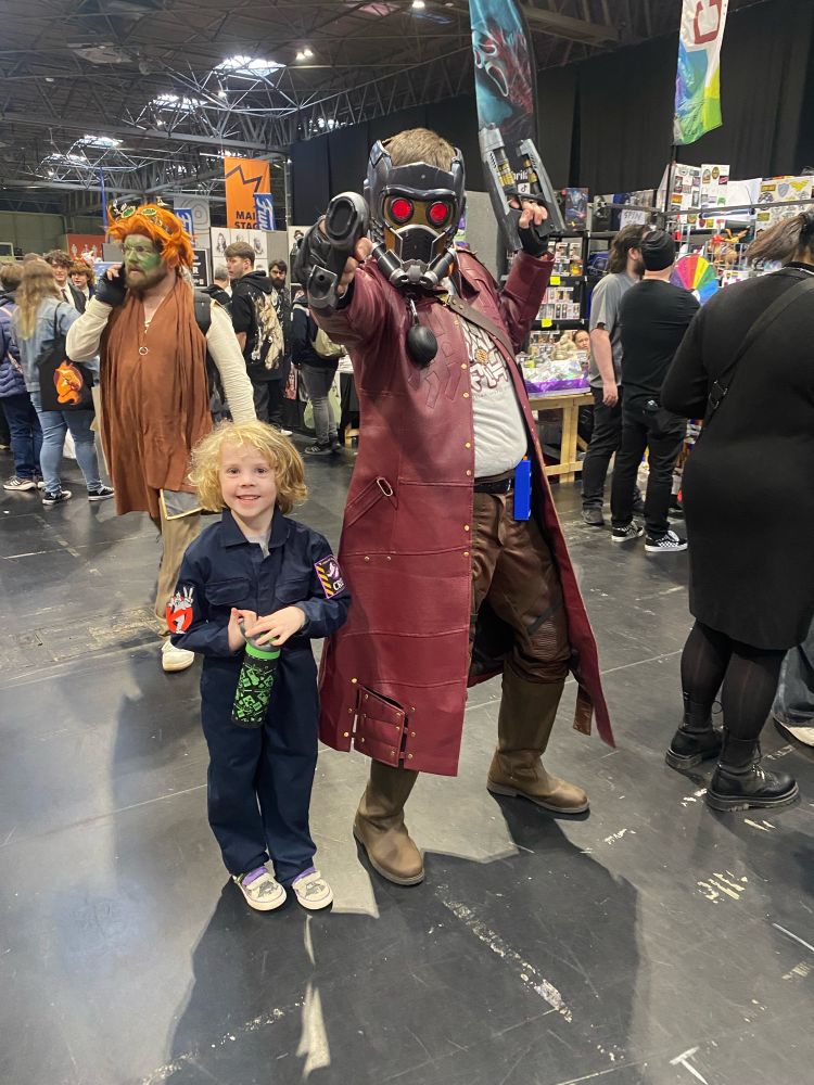 A beaming, blonde-haired child dressed as a Ghostbuster standing next to an adult in a good Star-Lord costume. They are at Comic-Con