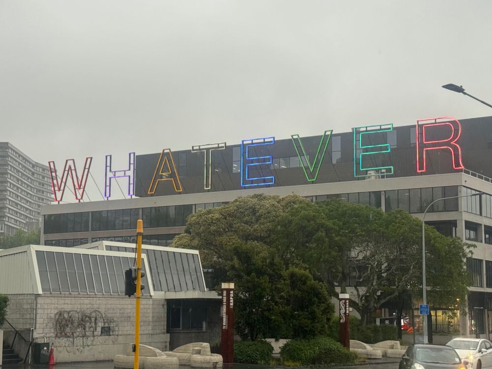 A neon sign, each letter a different colour, spelling "WHATEVER", mounted on top of an office building, seen through spring rain. There is a large tree just in front of the neon sign. 
