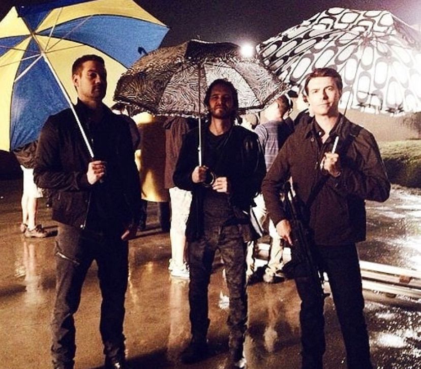 A behind-the-scenes photo from The CW’s Nikita. Pictured from left to right: Shane West, Aaron Stanford, and Noah Bean. They’re all holding silly umbrellas. 