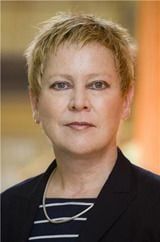 Headshot of a white woman with short blonde hair, wearing a black striped shirt, blazer, and necklace.