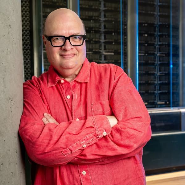 A bald white man wearing black rectangular glasses leans against a wall. He is wearing a red linen button-down shirt. His arms are crossed and he is smiling mischieviously.