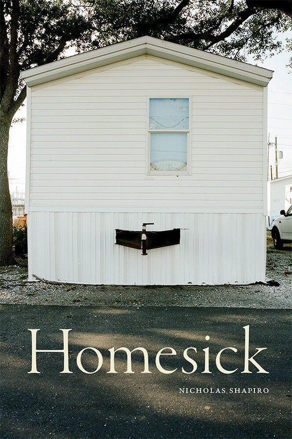 Cover of Homesick by Nicholas Shapiro. A small white mobile home is at the center of cover image, with a single window and a pipe protruding from its base. The scene is quiet and minimal, with gravel and asphalt in the foreground and large trees framing the background. The title “Homesick” appears in large, minimalistic serif text across the bottom, with the author’s name in smaller capital letters beneath it.