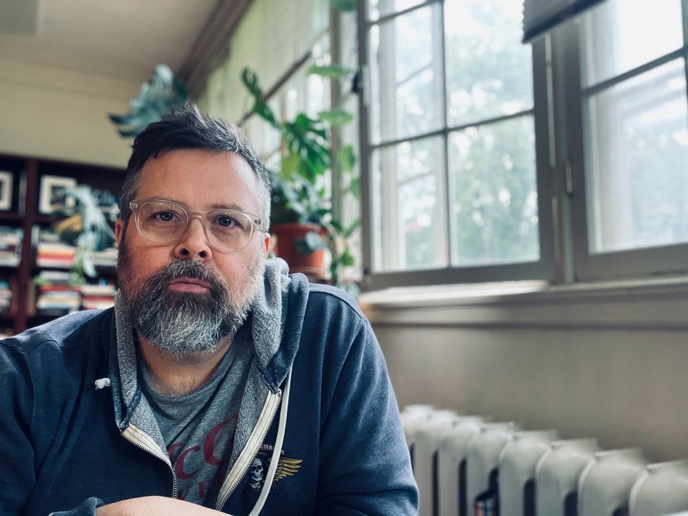 A white man with glasses and short gray hair and a gray beard sits in front of a radiator. He is wearing a grey hoodie. There is a plant over his shoulder.