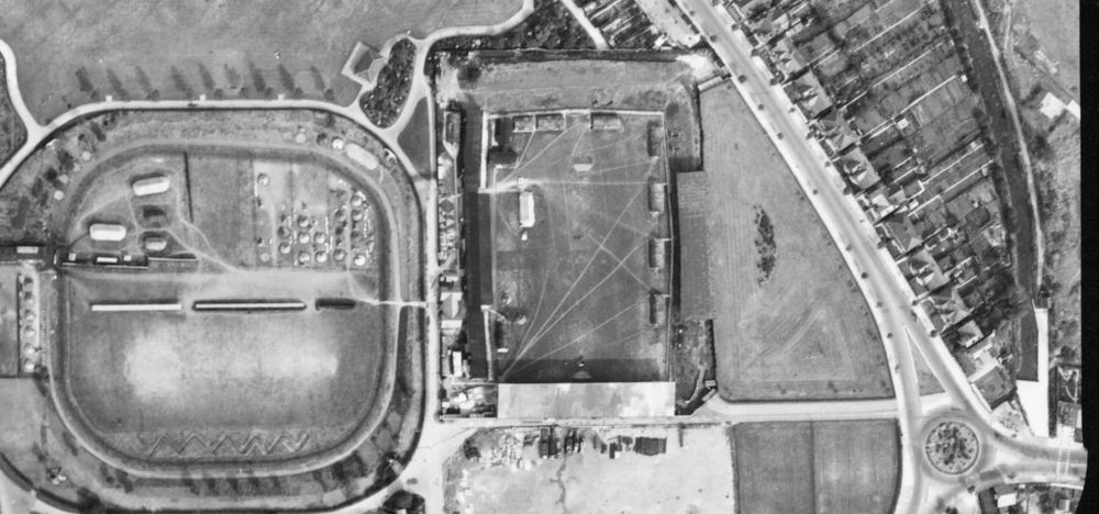 A black and white aerial photograph of the County Ground football and cricket pitches with tents on, with mud and well-worn paths visible. A single roundabout where the Magic Roundabout stands today.
Photograph from a USAAF flight, 8th September 1943, via Historica England.