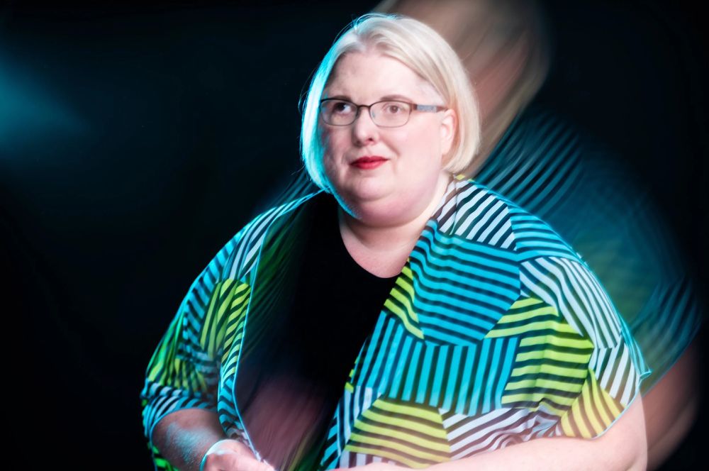 Ariel baska. A caucasion person with chin length light hair, glassed and a crooked smile. They wear a black shirt with a layered patchwork of blue and green stripes atop. The image carries a blur effect adding a mysterious glow to them.