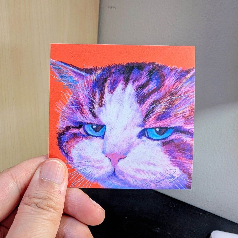 Photo of a left hand holding a square sticker. The sticker is a matte painting of a disappointed cat.