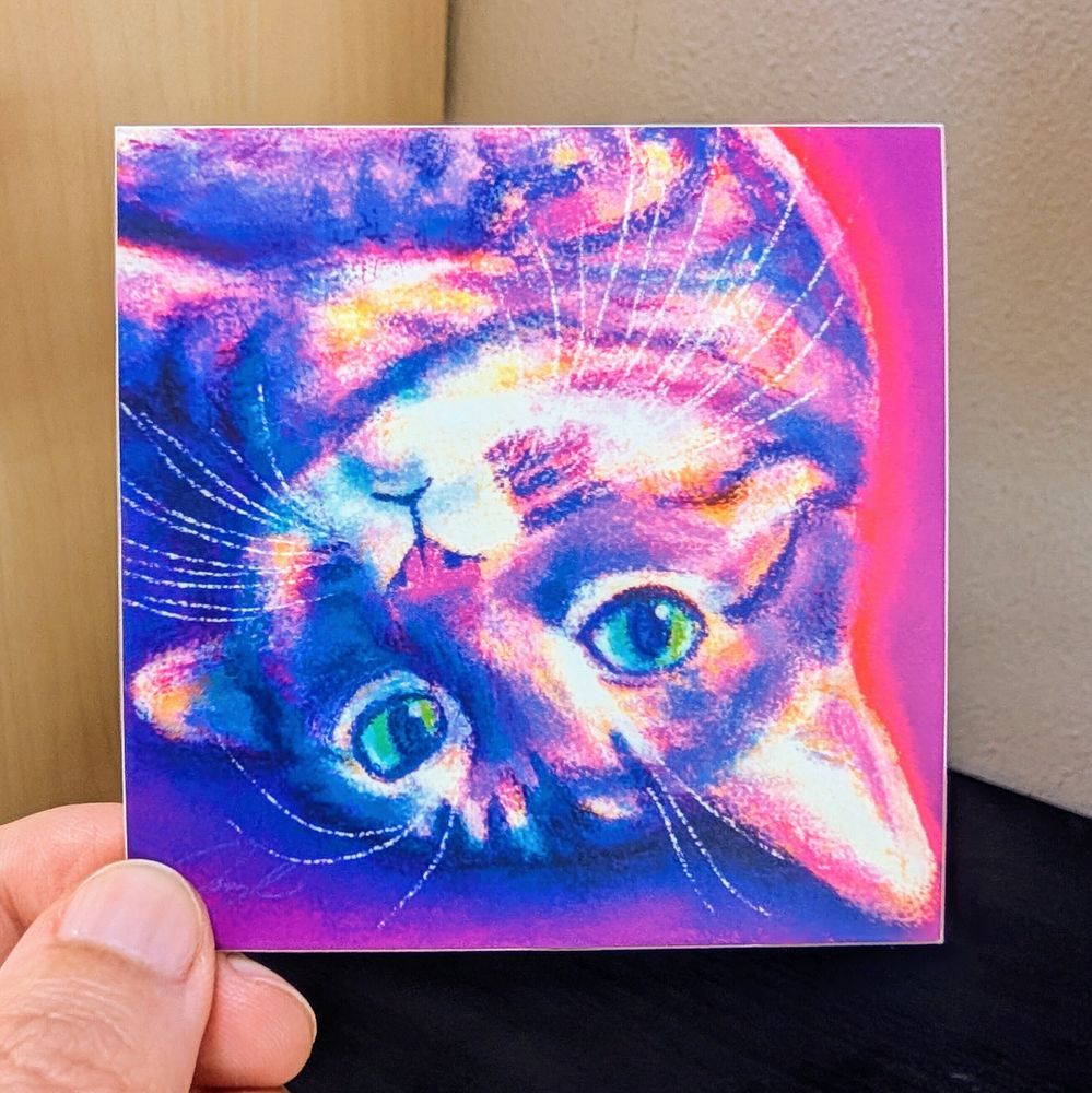 Photo of a left hand holding a square sticker. The sticker is a matte painting of a cat flopped on the ground looking up.