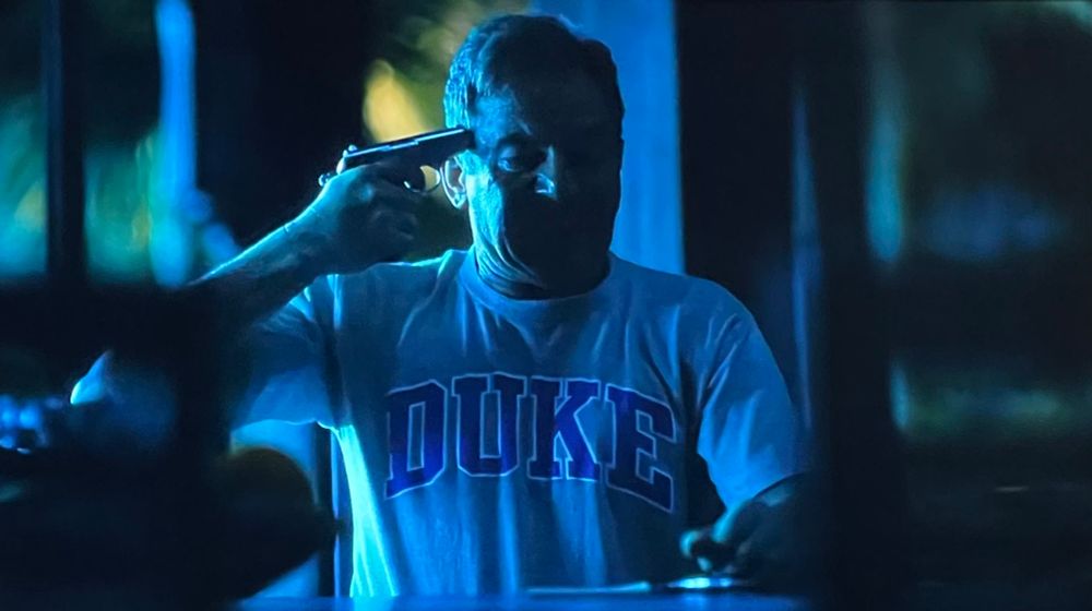 A man in Duke University T-shirt holds a gun to his head. I believe it’s from the show White Lotus 