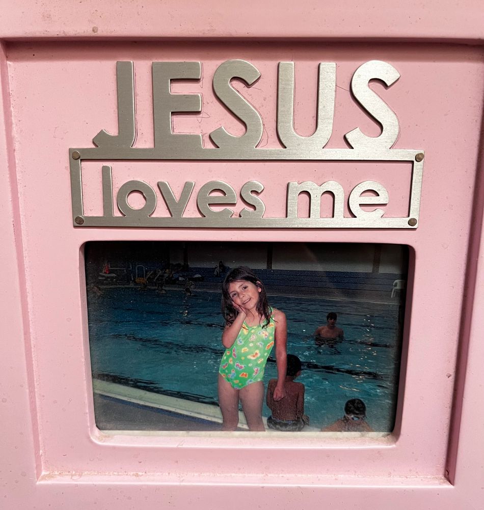 A pink photo frame that says “Jesus loves me” with a photo of 5-6 year old me in it