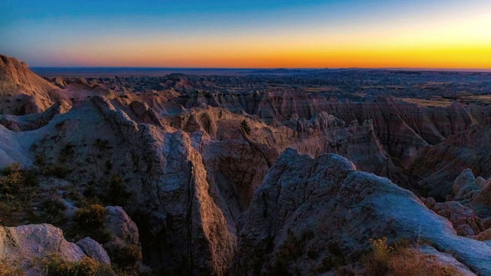 This panoramic photo captures a sunset over the dramatic, rugged landscape of the Badlands in South Dakota.

The image is split horizontally between the land and the sky, each with its own captivating color palette. In the foreground and stretching to the horizon, the Badlands' signature formations dominate the scene. They are a jumble of steep, sculpted hills, deep ravines, and eroded spires. The setting sun casts a warm, golden light across the landscape, illuminating the textures and layers of the rock formations and highlighting the stark contrast between light and shadow. The colors of the rocks range from soft grays and tans to hints of brown and rust.

Above the horizon, the sky transitions through a beautiful gradient. The lower part is a vibrant orange and yellow, a clear reflection of the sun as it dips below the horizon. As the eye moves upward, the colors fade into a soft blue, creating a peaceful and serene backdrop to the jagged terrain below. The meeting of the warm, earthy tones of the Badlands and the cool, vivid colors of the sky creates a powerful visual contrast, showcasing the unique beauty of this natural wonder at dusk.
