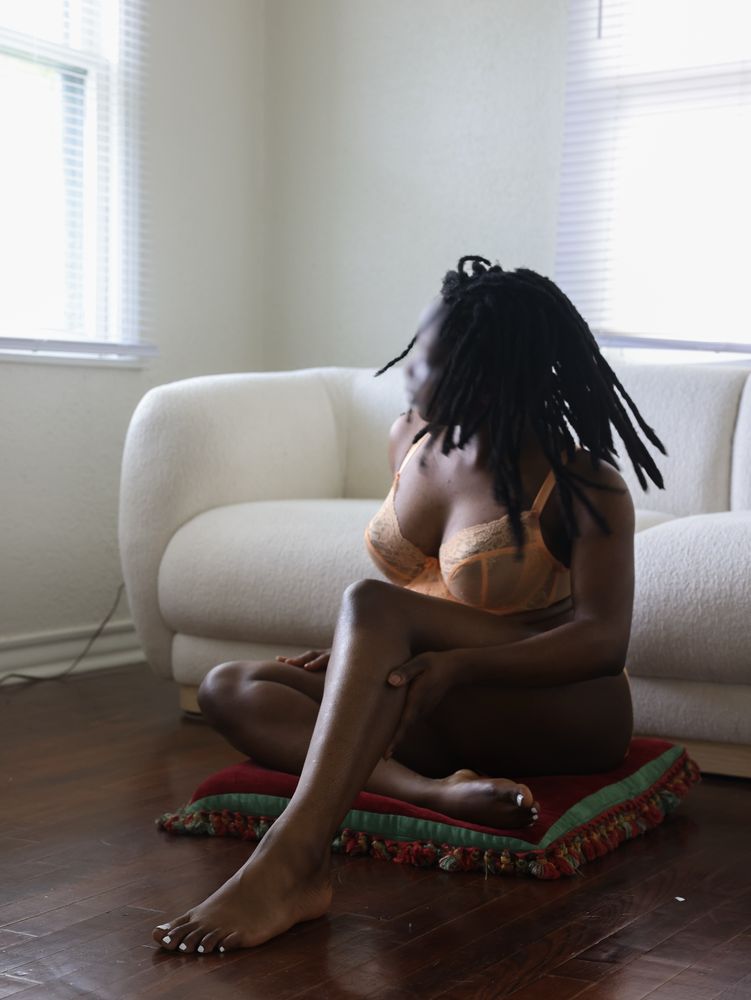 Black fem in pastel orange lingerie sitting on a maroon and green down pillow in front of a cream-colored Japandi style couch. One leg is crossed inward and the other is bent forward