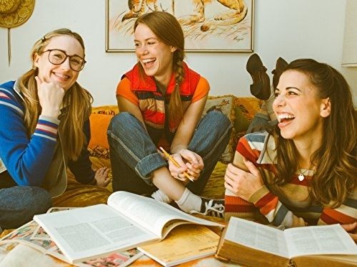 Photograph from critical role's retro photoshoot, in which, left to right, Ashley Johnson, marisha Ray, and Laura Bailey are casually hanging out in '70s style color blocked clothing. Ashley has nerd glasses on, Marisha is laughing while sitting cross-legged, and Laura is lying on her stomach. Can see her feet crossed behind her. 