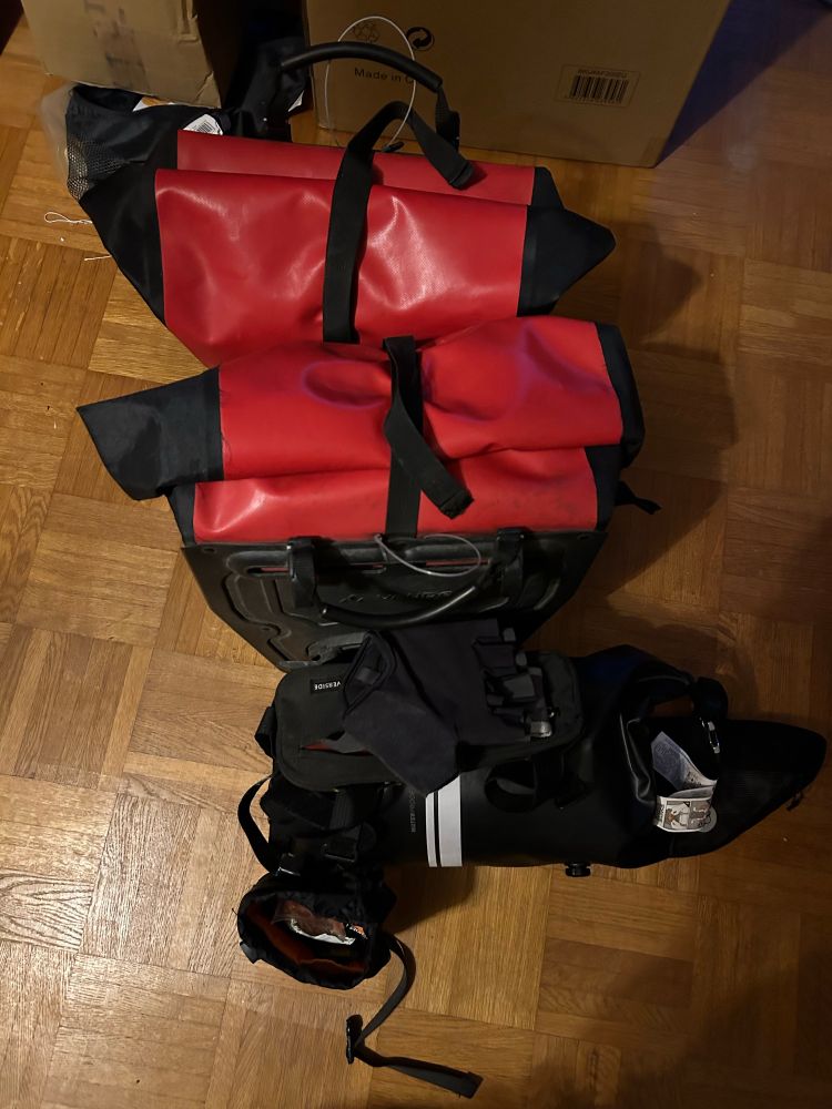 Zwei rote Satteltaschen, eine mittlere Rahmentasche, auf der eine Lenkerrolle sowie eine kleine Tasche für das Rahmenrohr liegen und ein Snackpouch, alles von oben auf Stäbchenparkett fotografiert. Alle kleinen Taschen sind schwarz.