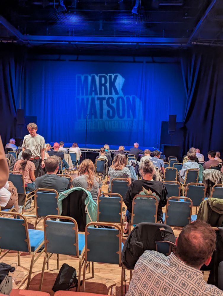 The stage is set. The set in question is a projection of the words Mark Watson on the back drapes.