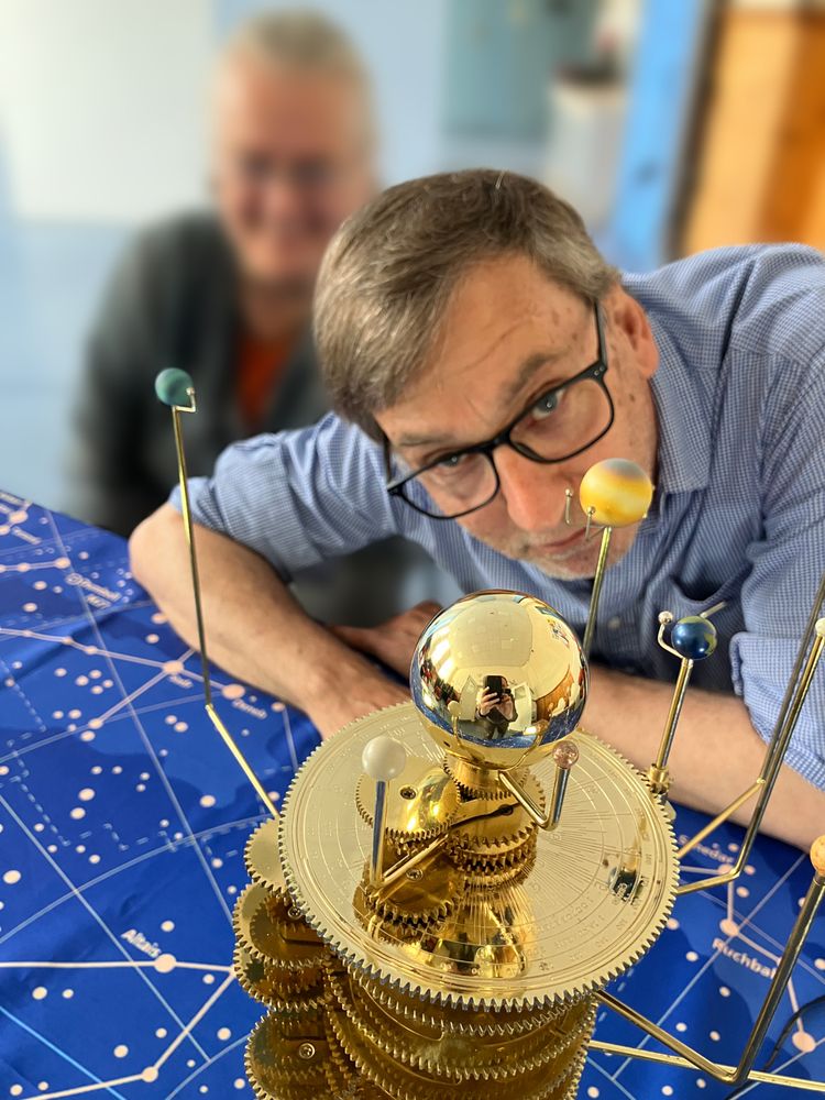In the foreground, a brass Orrery with lots of cogs and model planets showing. The orrery sits on a cloth with constellations and stars marked. Behind the orrery are two people, the closer wearing glasses, and further one who is too out-of-focus to see clearly.