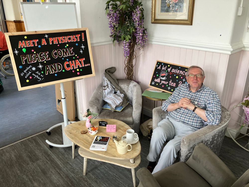 A table and three chairs in the corner of a cafe. One of the chairs is occupied by physicist Harold. Two blackboard signs behind the table read “Meet a Physicist! Please come and chat…” and “A bit of science with your cup of tea?”