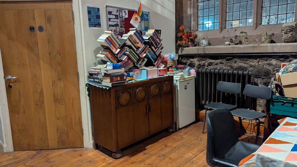 The western part of the north aisle of St nicks showing a toilet door, a cupboard topped by piles of books, a noticeboard, and a mini fridge 